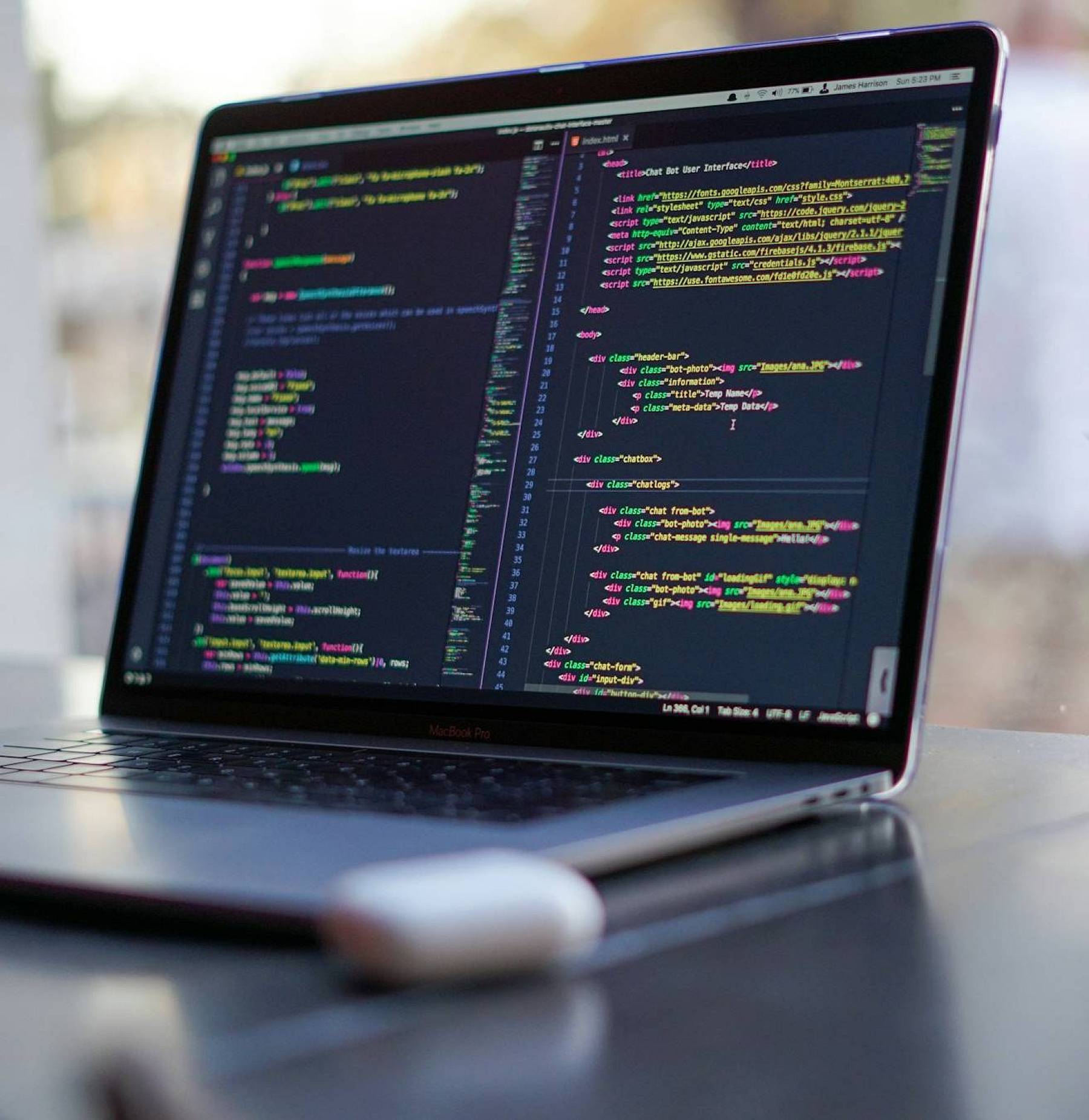 Ein aufgeklappter Laptop steht auf einem aufgeräumten, modernen schwarzen Schreibtisch. Auf dem Display des Laptops sind Codezeilen in einem Codingprogramm zu sehen.